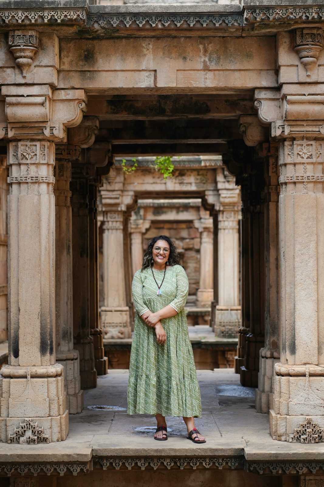 Ashmi Patel standing in an ancient carved stone stepwell, smiling, in a sage green dress